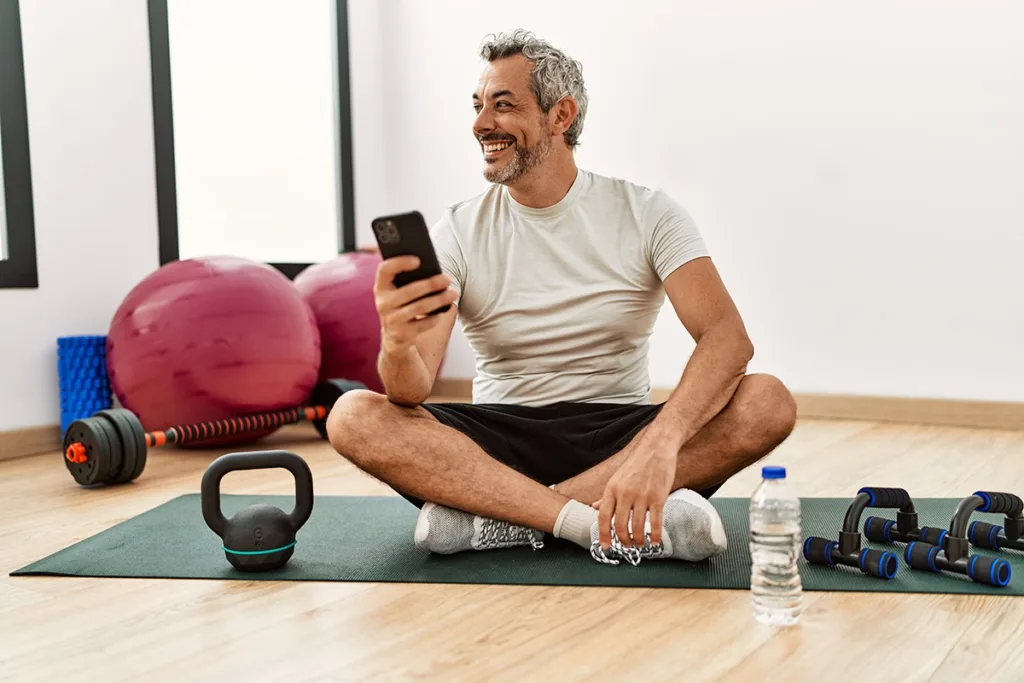 A middle aged man wearing an AI hearing aid at the gym with a yoga mat and dumbells, happily using an AI hearing aid app
